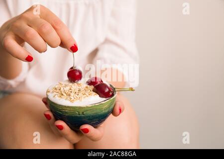 Nahaufnahme des Woman's Hände, die Schüssel mit organischen hausgemachte Joghurt mit Haferflocken und Kirschen. Hände mit Tasse vanille Joghurt mit Müsli Stockfoto