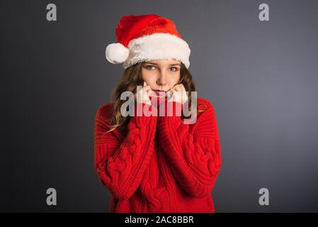 Frohe Weihnachten Schönheit Frau trägt rotes Hemd auf grauem Hintergrund. Ein nettes Mädchen in einem roten großen Pullover hält ihre Hände um ihr Gesicht und lächelt. Die Stockfoto