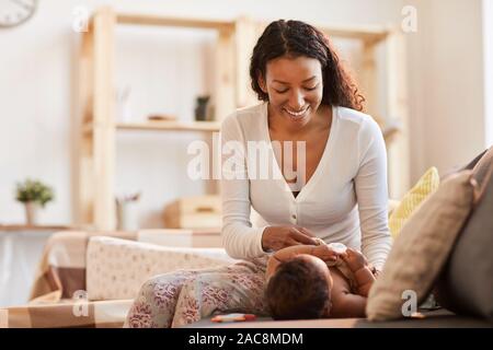 In warmen Farbtönen Porträt der Pflege Afro-amerikanische Mutter Dressing ihr Baby zu Hause auf der Couch, kopieren Raum Stockfoto