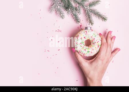 Weibliche Hand hängt Weihnachtskugel Dekoration aus Krapfen auf rosa Hintergrund. Weihnachten kreative Konzept. Stockfoto