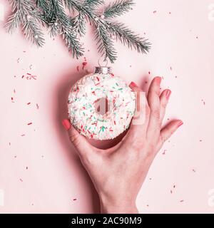 Weibliche Hand hängt Weihnachtskugel aus glasierten Krapfen auf rosa Hintergrund gemacht. Weihnachten kreative Konzept. Stockfoto