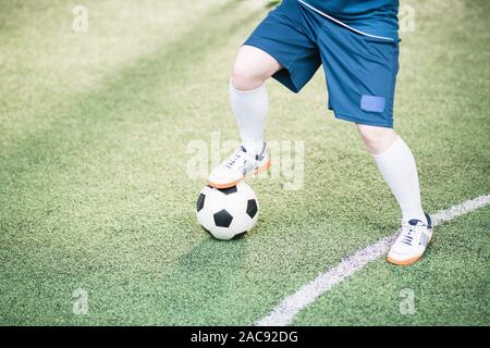 Beine der jungen aktiven, weiblichen Spieler in der blauen Uniform halten rechten Fuß auf Fußball während der Fußball auf dem Feld Stockfoto