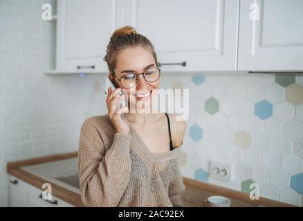 Schönen lächelnden jungen Frau fair lange Haare Mädchen tragen in der gemütlichen in Form gestrickter Pullover mit mobilen am hellen Küche Stockfoto