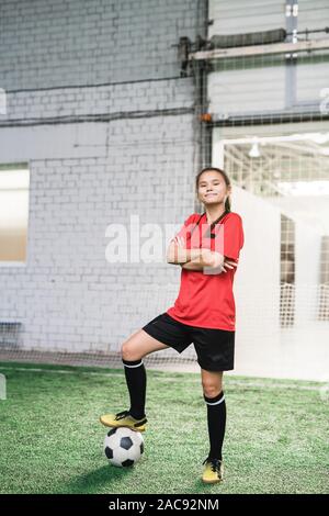 Glückliche junge erfolgreiche weibliche Fußball-Spieler im Sport einheitliche überqueren, um die Arme auf der Brust und halten für Fußball Stockfoto