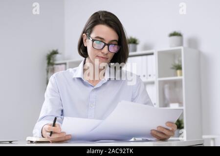 Junge ernsthafte weibliche Buchhalter in Brillen und formale Lesen finanzielle Beiträge von Arbeitsplatz Stockfoto