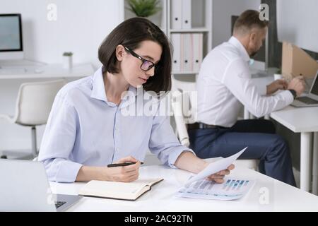 Besetzt weiblichen Office Manager durch Papiere und Notizen während der Sitzung von Schreibtisch mit Kollegen im Hintergrund Stockfoto