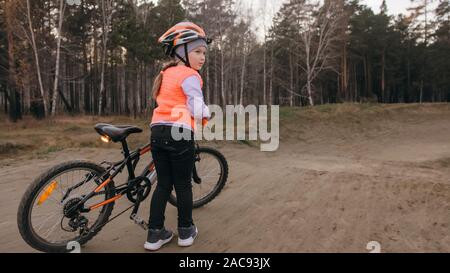 Eine kaukasische Kinder rides Bike Road Track in Dirt Park. Mädchen reiten schwarz orange Zyklus in der Rennstrecke. Kid geht Fahrrad Sport tun. Biker Motion Ride Stockfoto