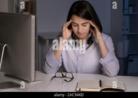 Junge müde Kundendienstmitarbeiter Kopf berühren, während Sie von Arbeitsplatz sitzen am späten Abend Stockfoto