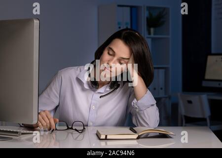 Müde weibliche hotline Betreiber mit geschlossenen Augen napping von Schreibtisch, während spät am Abend im dunklen Büro arbeiten Stockfoto