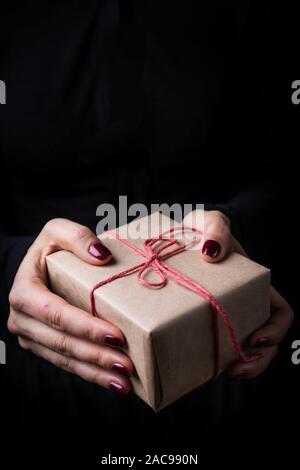 Nahaufnahme von weibliche Hände halten ein kleines Geschenk mit roter Schleife verpackt. Kleines Geschenk in den Händen einer Frau auf schwarzen Hintergrund. Stockfoto