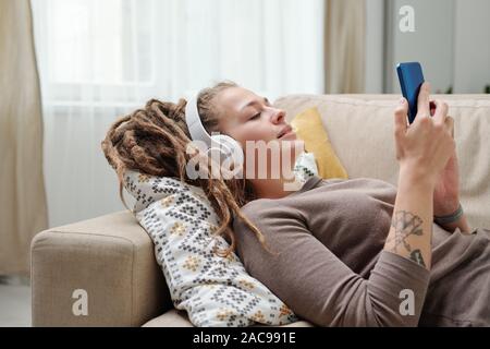 Hübsche junge ruhige Frau mit Kopfhörer Scrollen in Smartphone auf der Couch Stockfoto