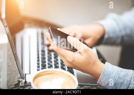 Nahaufnahme der Laptop auf dem Tisch. Mädchen in beige Pullover bei Tisch holding Smartphone sitzen. Frau entscheidet, Produkte im Online Store verwendet Computer. Eine Frau prüft die E-Mails beim Sitzen in Cafés. Stockfoto