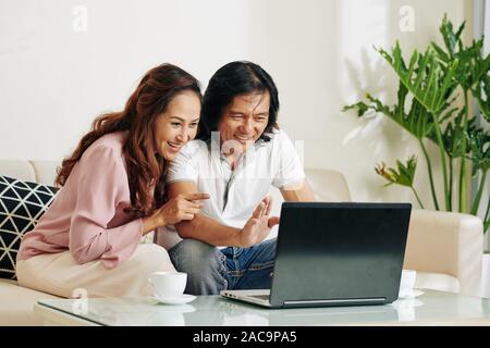 Fröhlich lachend im mittleren Alter asiatischen Paar video ihre Verwandten Anrufen beim Sitzen auf einem Sofa mit geöffneten Laptop Stockfoto