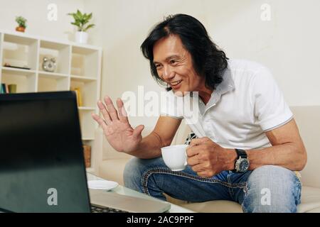 Gut gelaunt im mittleren Alter asiatischer Mann trinken Tasse Kaffee, wenn mit seinem Freund telefonieren über Laptop zu Hause Stockfoto