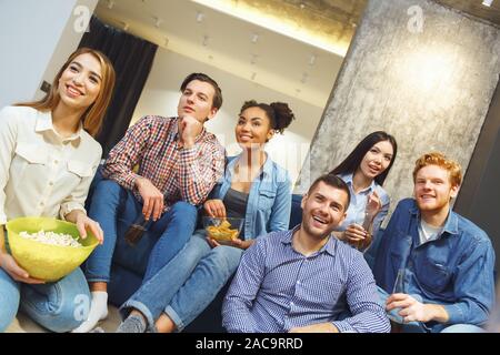 Junge Männer und Frauen sich gemeinsam amüsiert drinnen essen Snacks chips Fernsehen American football Meisterschaft lächelt Fröhlich Stockfoto