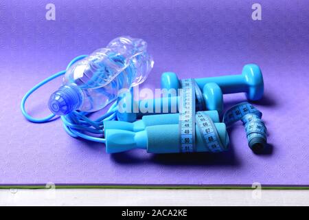 Sport und eine gesunde Lebensweise. Kurzhanteln, Wasserflasche und Seil springen in Cyan Farbe auf lila Hintergrund. Hanteln und Springseil mit Maßband Rollen gebunden. Gestaltung und Fitnessgeräte. Stockfoto