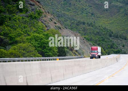 Red Big Rig Semi Truck mit Anhänger bergauf auf kurvenreichen in Mountain pass Highway mit einem anderen Lkw und Pkw Verkehr und grüne Wälder auf backgro Stockfoto