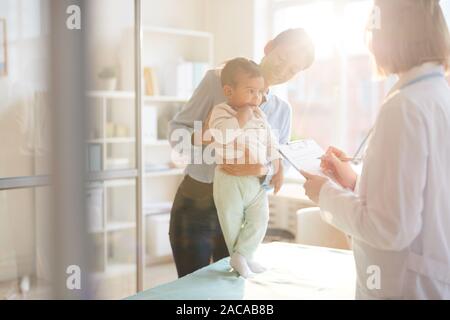 Ärztin im weißen Kittel stehen und Schreiben einer Behandlung, während die Mutter, die ihr Baby während ihres Besuchs im Krankenhaus Stockfoto