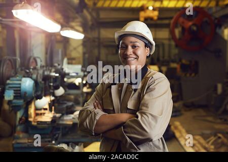 Taille bis Portrait von Mixed-race Arbeiterin selbstbewusst posiert im Stehen mit der Waffe in der Werkhalle gekreuzt Stockfoto