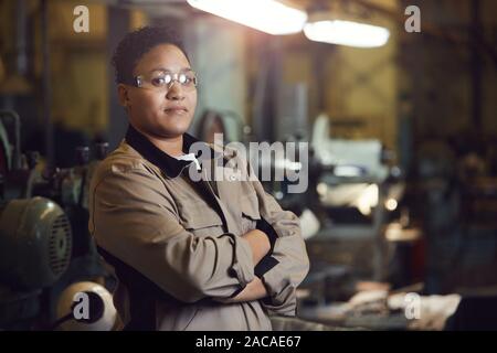 Taille bis Portrait von Mixed-race Arbeiterin selbstbewusst posiert im Stehen mit den Armen in der Werkstatt gekreuzt Stockfoto