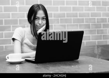 Junge schöne asiatische Jugendmädchen ruhenden Kinn auf Hand mit Laptop und Cappuccino auf hölzernen Tisch gegen die Wand Stockfoto