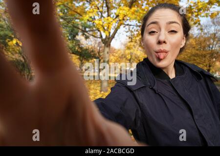 Freche junge Frau in ihrem 20s die Zunge heraus klemmt unter selfie Foto im Park mit Herbstfarben posing - Wide Angle Shot Holding unsichtbare Smartphone mit ausgestrecktem Arm Stockfoto