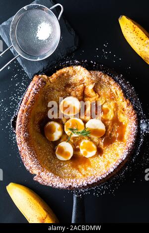 Essen Konzept hausgemachte Niederländische Baby oder Deutsche Banane caramel topping Pfannkuchen in der Pfanne Eisen auf schwarzem Hintergrund cast Stockfoto