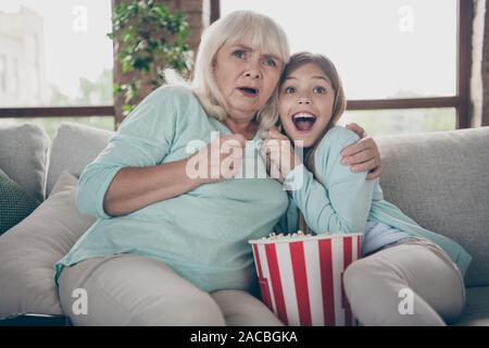 Foto von zwei Menschen weiß behaarte Oma kleine Enkelin sitzen Sofa fernsehen Popcorn scary Horror Film Sommer Wochenende zusammen essen Haus Stockfoto