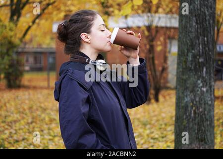 Trendige junge Frau hipster tragen parka Mantel mit Kopfhörer um den Hals und das Trinken von Kaffee Tasse zu beim Gehen im Park an einem kalten Herbsttag. Stockfoto