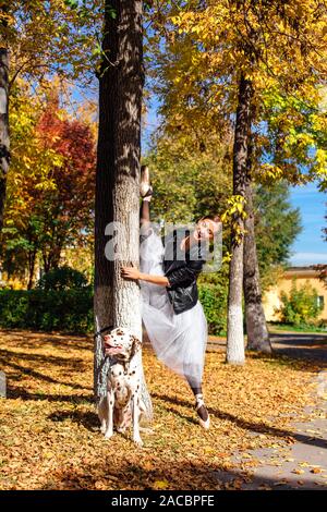 Ballerina mit Dalmatiner Hund im Park. Frau Ballerina in einem weißen Ballett Rock und schwarze Lederjacke, Splits in Pointe Shoes im Herbst Park Stockfoto