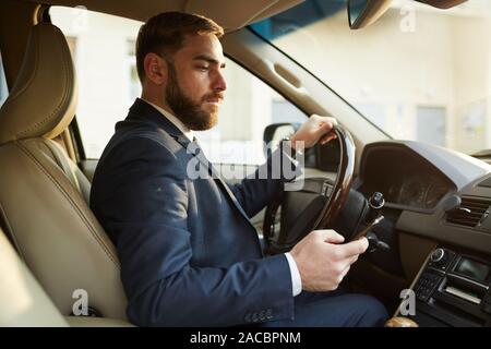 Junge Unternehmer, die sich hinter dem Lenkrad sitzen und Fahren, während dem Lesen einer Nachricht auf seinem Handy Stockfoto