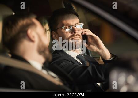 Ernsthafte bärtigen Mann in Brillen Gespräch am Handy und mit Blick auf die Passagier während ihrer Reise auf dem Auto Stockfoto