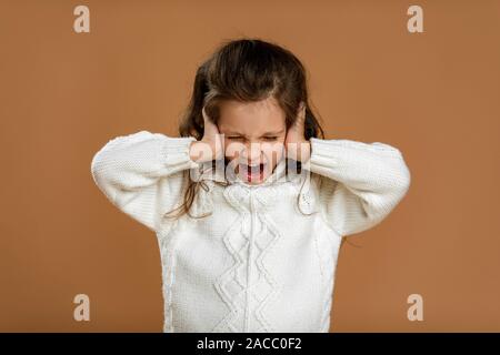 Cute curly kleines Mädchen in weißen Pullover schließt ihre Ohren mit ihrer Hand und schreien auf beigem Hintergrund. Stockfoto