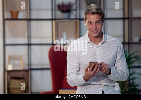 Lächelnder junger Mann in kabellosen Kopfhörern, Portrait im Loft-Interieur, stilvolles Zimmer. Positiver Kerl verwendet Smartphone für Online-Schulungen. Stockfoto