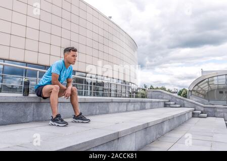 Männliche Athlet sitzt auf der Treppe und schaut in die Ferne, liegt nach dem Workout und genießt, Training Training im Sommer Stadt. Fitness motivation Jugend Stockfoto