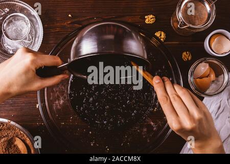 Der Teig für Schokoladenkuchen Kuchen, die Hände des Weiblichen gießen geschmolzene Schokolade mit Walnüssen in Schale auf Holztisch unter Zutaten und Utensilien Stockfoto