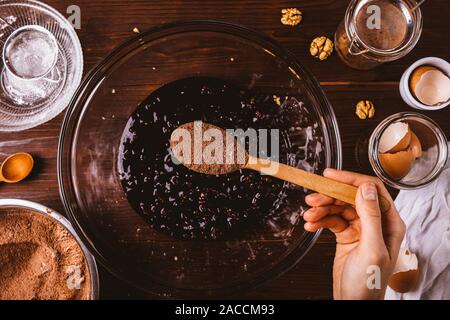 Weibliche Hand hinzufügen Esslöffel Kakaopulver in geschmolzene Schokolade mit Walnüssen Teig für köstliche, hausgemachte Kuchen, Brownies, Ansicht von oben. Stockfoto