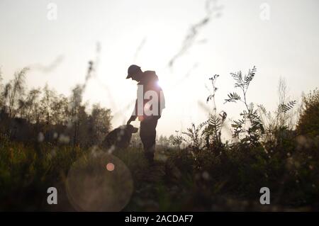 Silhouette eines Mannes mit einem Hund auf einem Spaziergang. Border Collie auf der Wiese gegen die Strahlen der untergehenden Sonne. Stockfoto