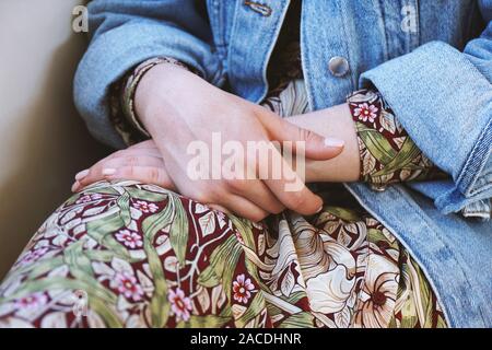 Den mittleren Abschnitt der jungen Frau das Tragen von Jeans Jacke über Sommer Kleid mit Blumenmuster - in der Nähe der weiblichen Hände in ihrem Schoß gefaltet Stockfoto
