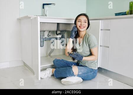 Stolz fröhliche junge asiatische Frau sitzt auf der Küche Fußboden und am Telefon sprechen nach der Fixierung tropfende Spüle Leitung Stockfoto
