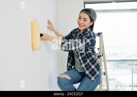 Glückliche junge asiatische Frau in Gap und Plaid Shirt Grundierung vor dem Auftragen von Putz an der Wand Stockfoto