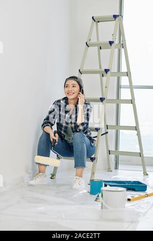 Fröhliche junge vietnamesische Frau sitzt auf der Treppe und ruft am Telefon nach dem Lackieren Zimmer in Ihre neue Wohnung Stockfoto