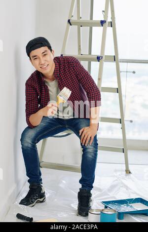 Positive junge vietnamse Mann sitzt auf Leiter mit Pinsel in der Hand und Suchen an Kamera Stockfoto