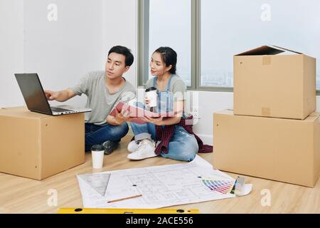 Junge vietnamesische Paare sitzen auf dem Boden in der neuen Wohnung mit dem Aufbau, der Planung und der Suche nach interior design Ideen online Stockfoto