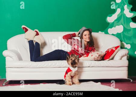 Junge Frau in Weihnachten Pullover liegen auf einem Sofa im Studio mit Weihnachtsschmuck. Yorkshire Terrier in Santa Kostüm Stockfoto