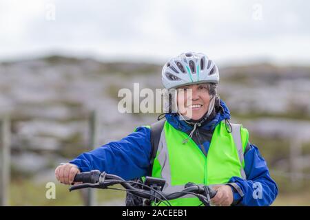 Lächelnd reife weibliche Radfahrer mit einer blauen Jacke, Helm und anti-reflektierende Weste neben einem Fahrrad mit einem unscharfen Hintergrund, entspannten Tag in Irland Stockfoto