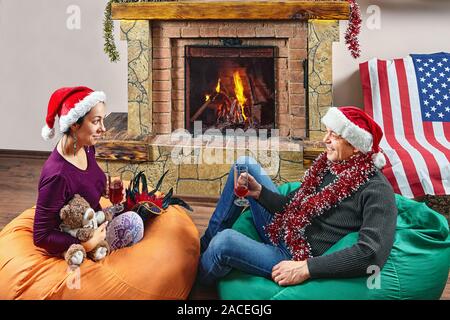 Frau und Mann in Santa's Hüte werden Wein trinken und auf Sitzsäcken beim feiern das neue Jahr. Ausgereifte Familie Paar feiert Weihnachten in der Nähe von firep Stockfoto