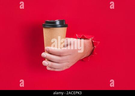 Männliche Hand Handwerk Papier Tasse Kaffee durch ein Loch in roten Hintergrund. Stockfoto