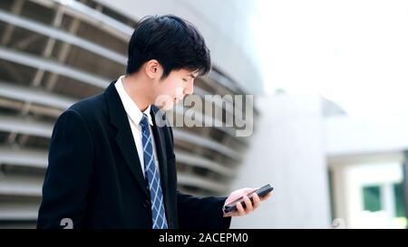 Junge Unternehmer mit mobilephone App texting außerhalb des Büros mit Wolkenkratzern Gebäude im Hintergrund. Junger Mann mit Smartphone für Business Stockfoto