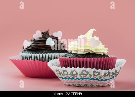 Chocolate Cupcake mit Vanille Schokolade Vereisung und essbar Herzen, in Schönes dekoratives Papier Schimmel, rosa Hintergrund Stockfoto